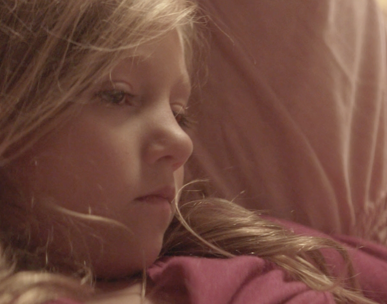 A young girl with long blonde hair lies on her side, gazing thoughtfully into the distance. Warm, soft lighting creates a calm and introspective atmosphere.