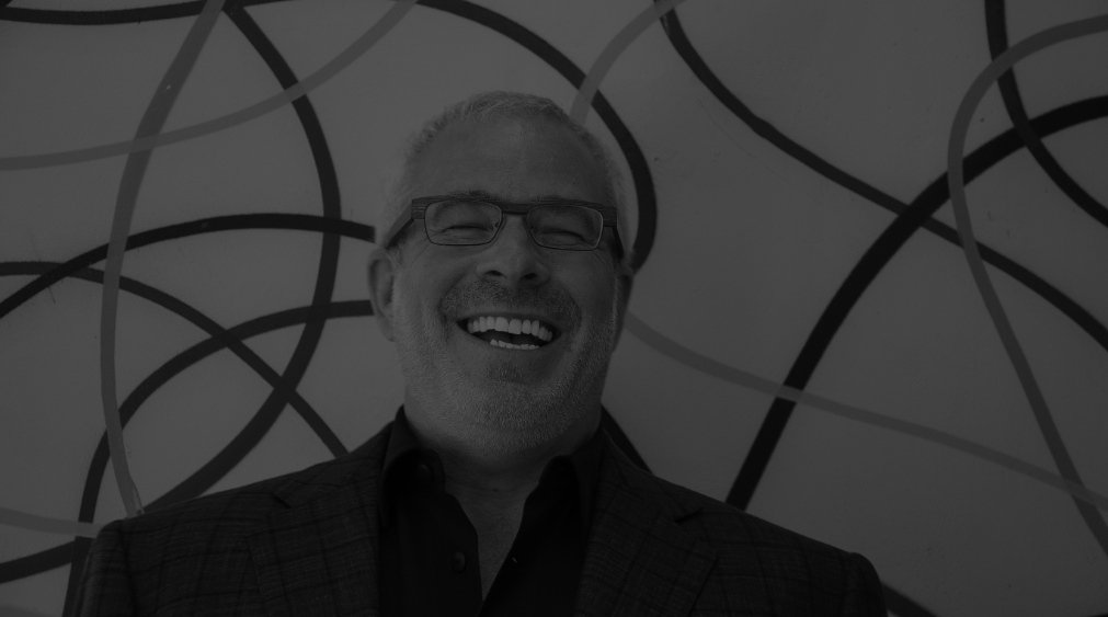 A man with short gray hair, glasses, and a beard is smiling and laughing. He is wearing a dark jacket and shirt, standing in front of a wall with a pattern of intersecting curved lines.