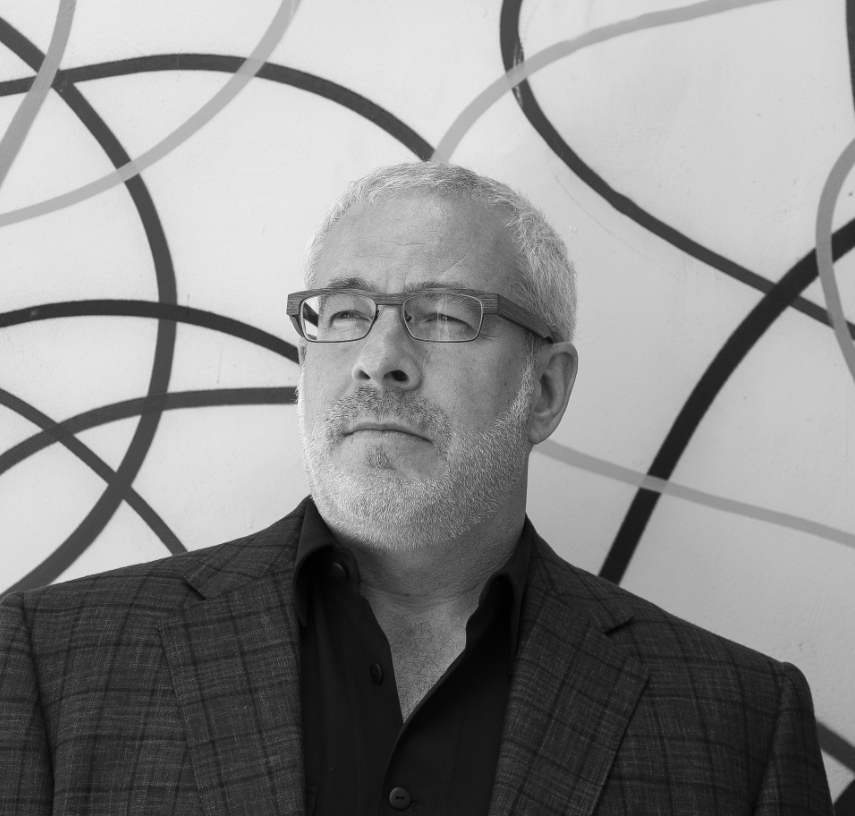 A man with short gray hair, glasses, and a trimmed beard wears a plaid jacket and dark shirt, standing in front of a white wall with abstract black and gray curved lines.