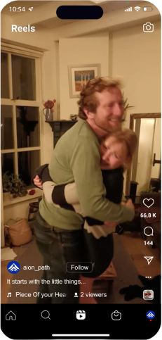 A man and woman smiling and hugging tightly in a warmly lit living room, with plants and a framed picture in the background. Instagram Reels interface elements and engagement icons are visible on the screen.