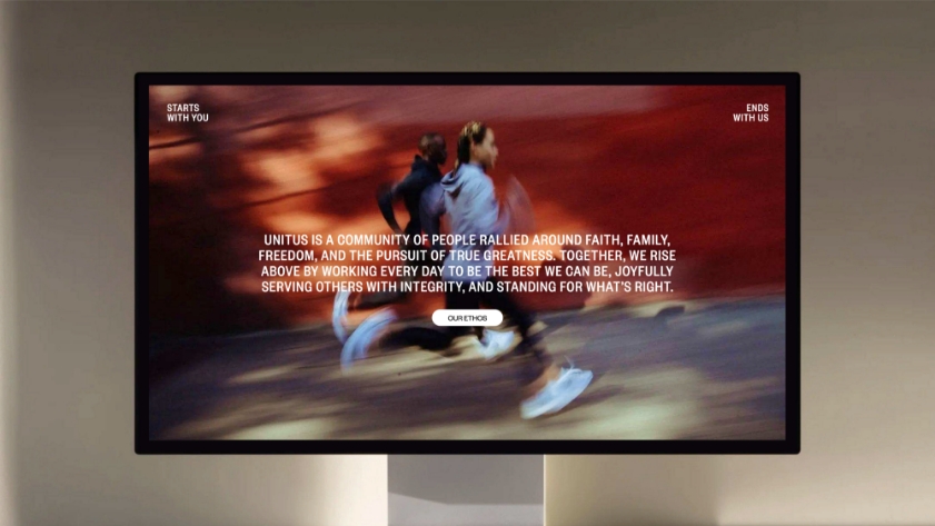 A computer monitor displays a blurred image of two people running, with motivational text overlay about community, greatness, and integrity. There is a button labeled OUR ETHOS in the center of the screen.