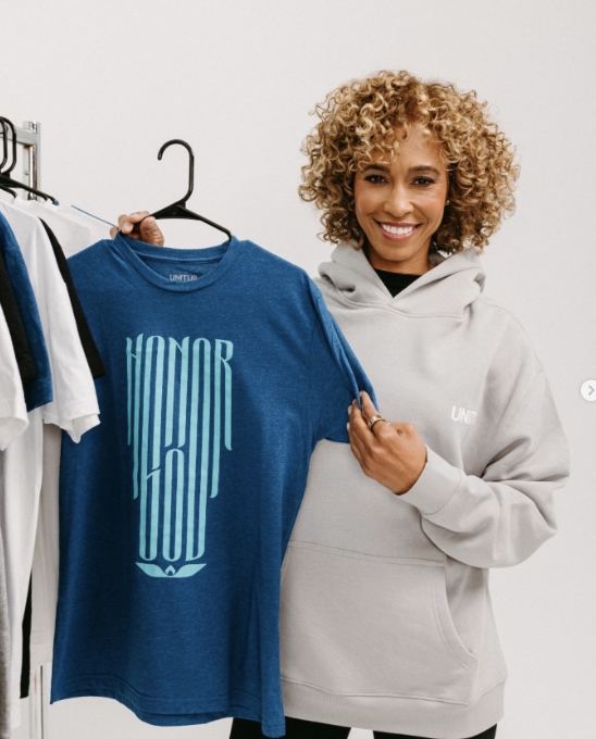 A smiling woman with curly hair wearing a light gray hoodie holds up a blue T-shirt that says HONOR GOD in bold letters. She stands next to a clothing rack with other shirts.