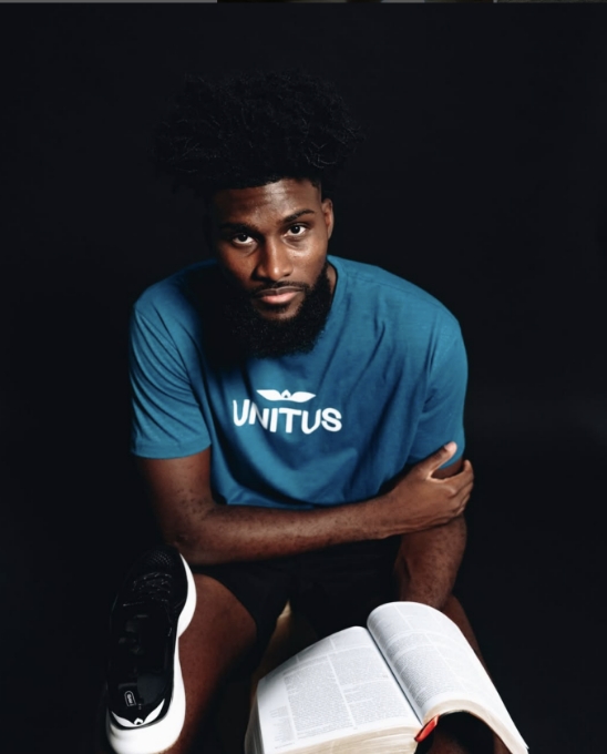 A man with a beard wearing a blue UNITUS t-shirt sits against a black background, looking at the camera. He holds an open book on his lap and rests one arm on his knee, with a sneaker visible in the lower corner.