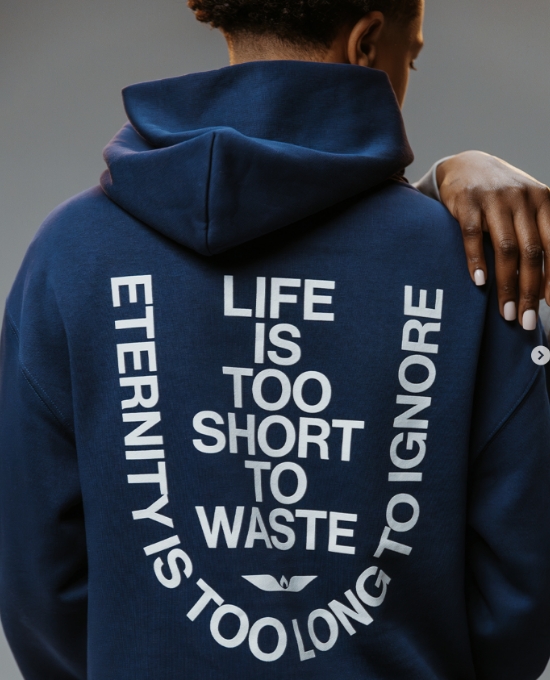 A person wearing a navy blue hoodie with the words Life is too short to waste, eternity is too long to ignore printed in bold white letters on the back. The person is facing away from the camera.