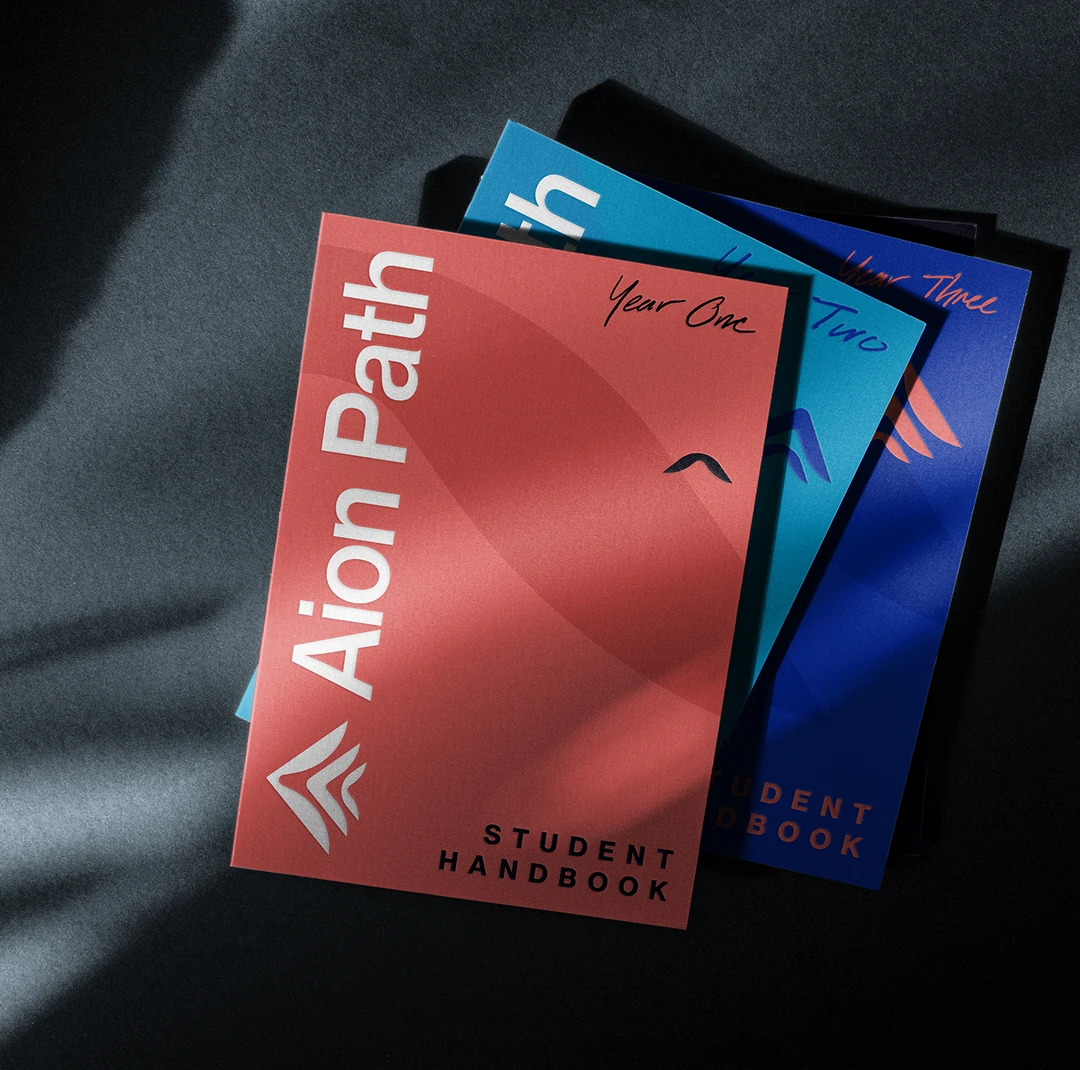 Three colorful student handbooks labeled Aion Path are stacked on a dark surface. The top one is red and says Year One, the second is teal and says Year Two, and the third is blue for Year Three.