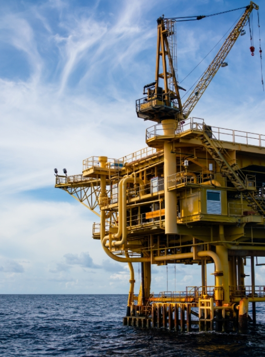 A yellow offshore oil rig stands above the ocean, with pipes and cranes visible, under a partly cloudy blue sky.