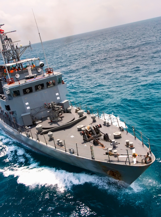 A military naval ship moves quickly through the ocean, creating white waves along its sides under a clear sky. The ships deck is equipped with antennas, radar, and other equipment.