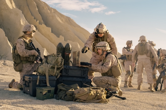 Several soldiers in desert camouflage uniforms are gathered around military gear and equipment cases in a sandy, rocky landscape; some are standing, while others kneel and work with the gear.
