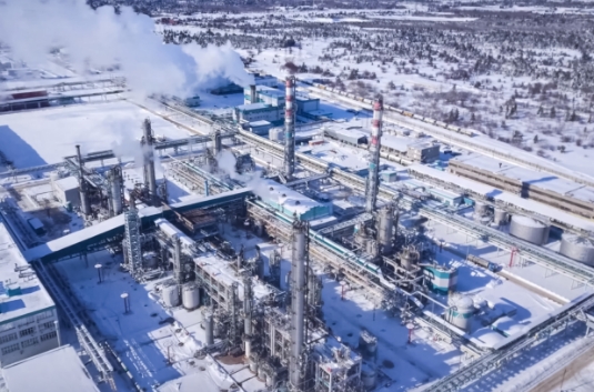 A large industrial facility with multiple smokestacks emitting steam, surrounded by snow-covered ground and trees, viewed from above in winter.