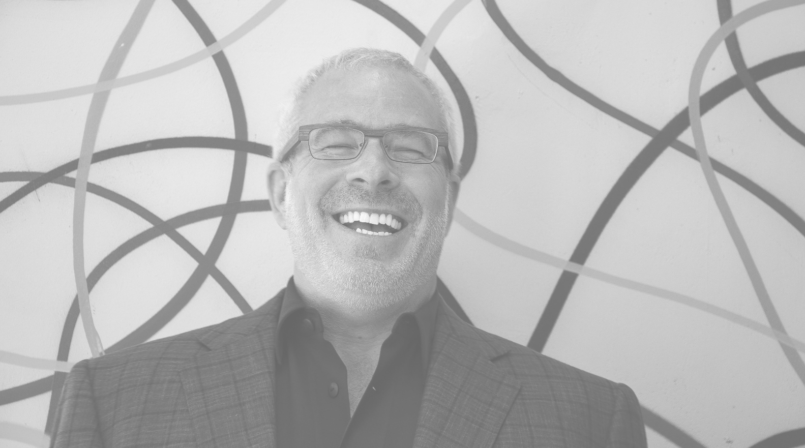 A man with short gray hair, glasses, and a beard is laughing while standing in front of a wall decorated with abstract, curved lines. He is wearing a dark shirt and a plaid blazer.