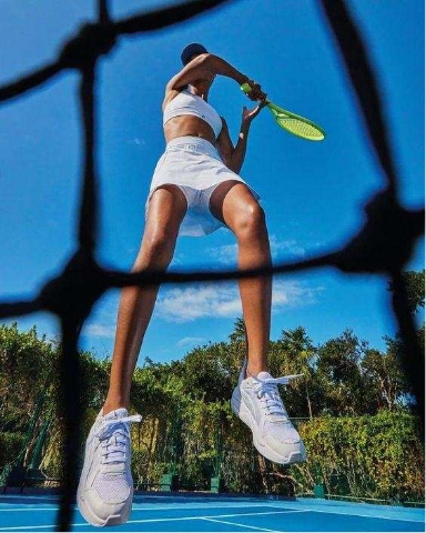 A tennis player in white sportswear and sneakers jumps to hit a shot with a yellow racket on an outdoor court, with a blue sky and greenery in the background, seen through the net from a low angle.