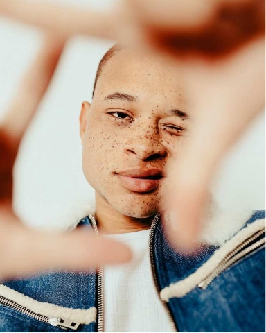 A person with short hair and freckles is winking at the camera while wearing a denim jacket. Their hands are in the foreground, forming a frame around their face. The background is plain and white.