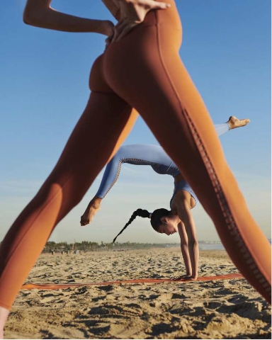 Two women practice yoga on a sandy beach. One, in orange leggings, stands with legs apart in the foreground, while the other, in blue leggings, does a handstand seen through the gap between the first womans legs.