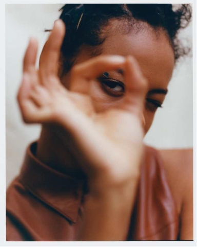 A person with curly hair gazes through their hand, which is forming a circle gesture in front of their face. The image is softly focused and the individual wears a brown sleeveless top.