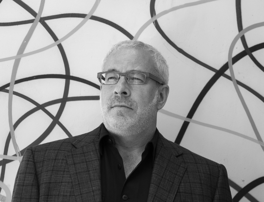 A man with short gray hair, glasses, and a beard wears a plaid jacket and dark shirt, standing in front of a white wall with abstract black and gray swirling lines.