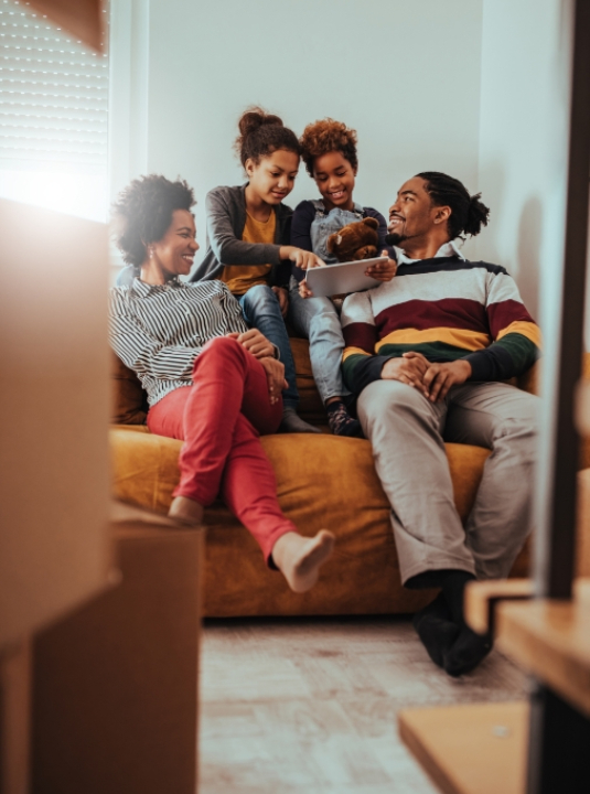 A smiling family of four sits on a couch at home, with two children playing on a tablet while the parents watch and laugh together, surrounded by moving boxes.