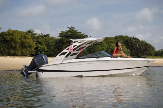 A woman in a red swimsuit sits on the bow of a white motorboat near a sandy shore, with trees in the background and an American flag attached to the boat.
