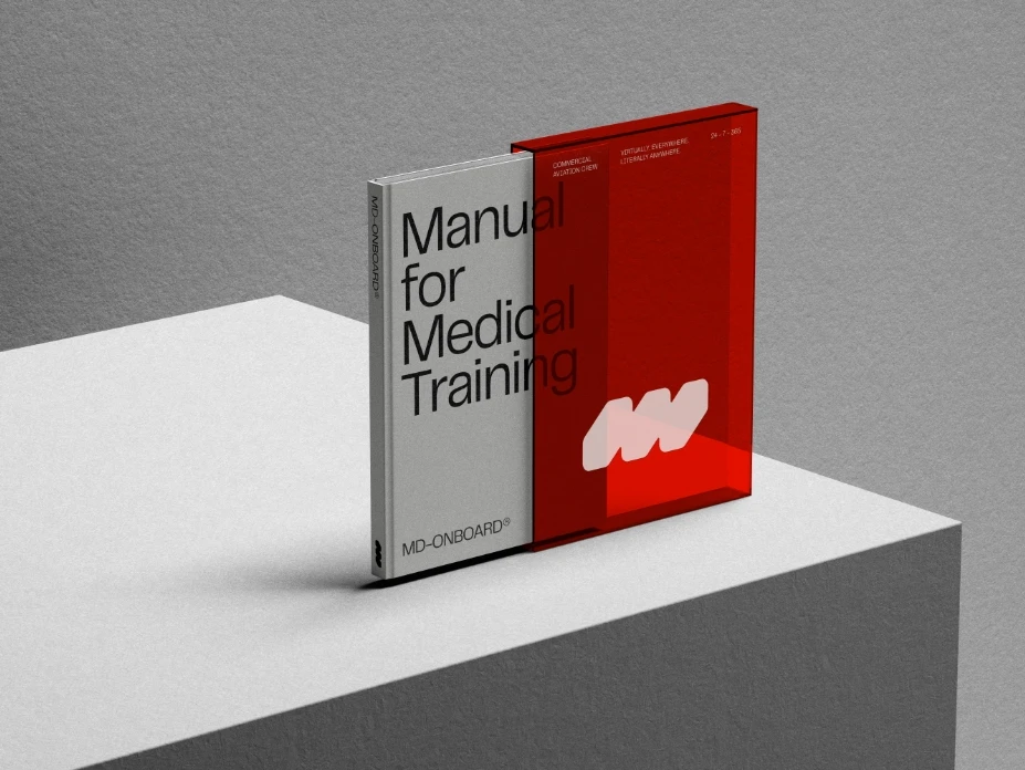 A book titled Manual for Medical Training with a minimalist white and black cover stands upright next to a translucent red slipcase displaying a bold white M logo, all placed on a geometric white pedestal.
