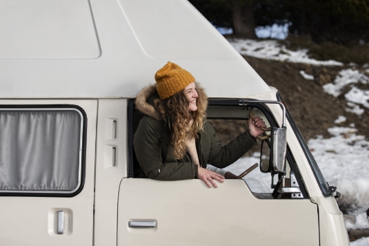 A woman wearing a yellow beanie and winter coat smiles while leaning out the window of a white camper van parked near a snowy area with trees in the background.