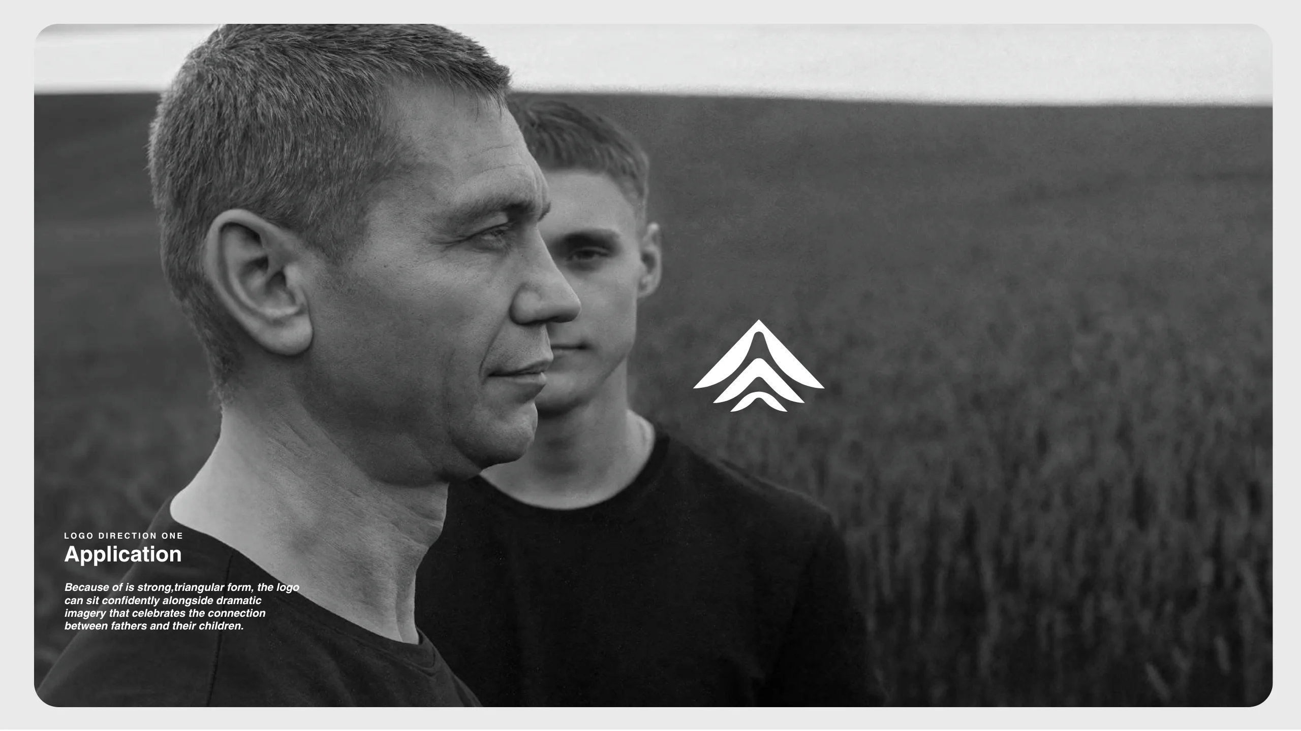 Two men stand outdoors facing slightly different directions, one closer to the camera. They are both wearing dark shirts. A minimalist triangular logo appears between them, and descriptive text is positioned on the left side of the image.