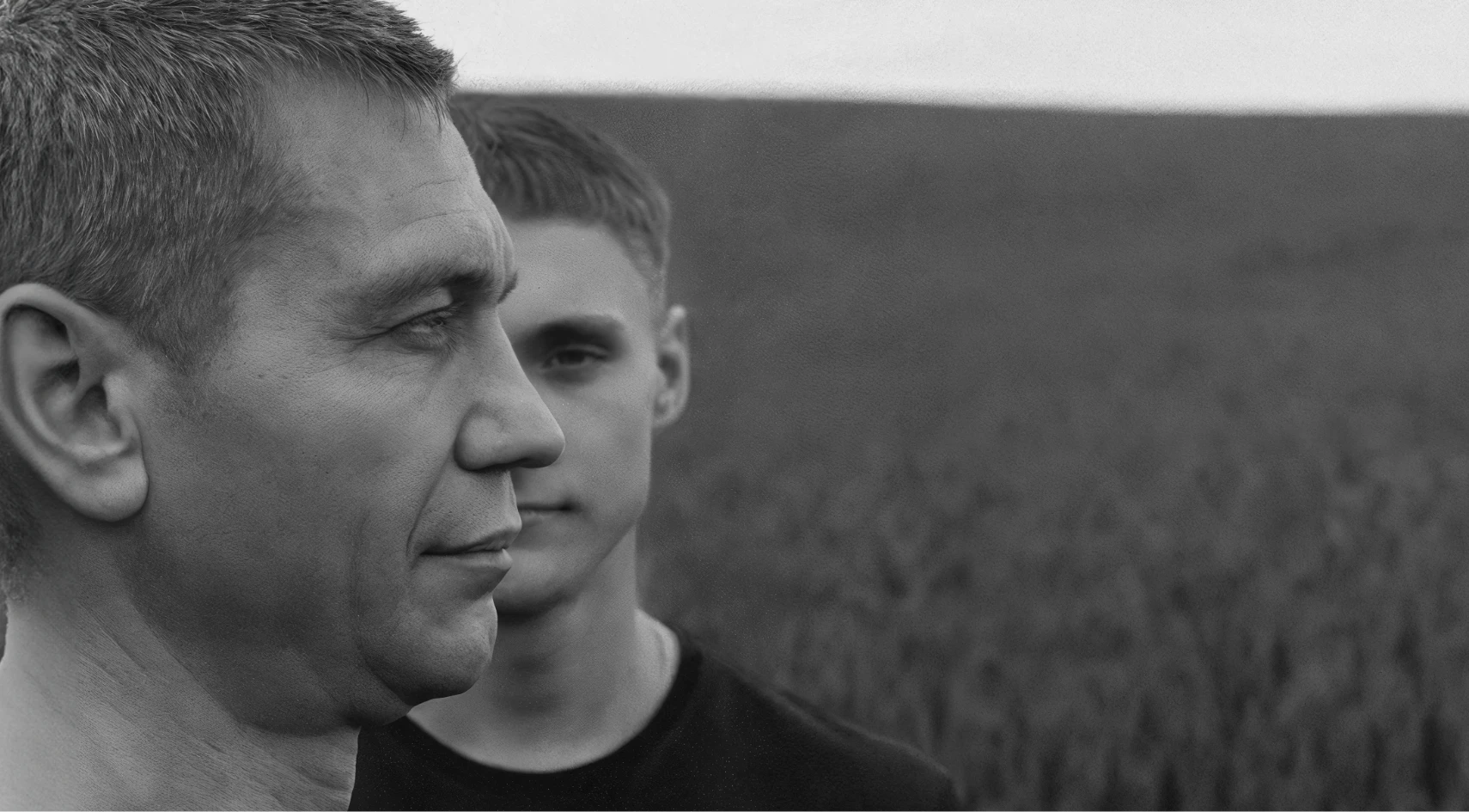 A black and white photo of two men standing in a field. The man in the foreground is in profile, facing left, while the younger man behind him looks forward. The background shows a blurred landscape.