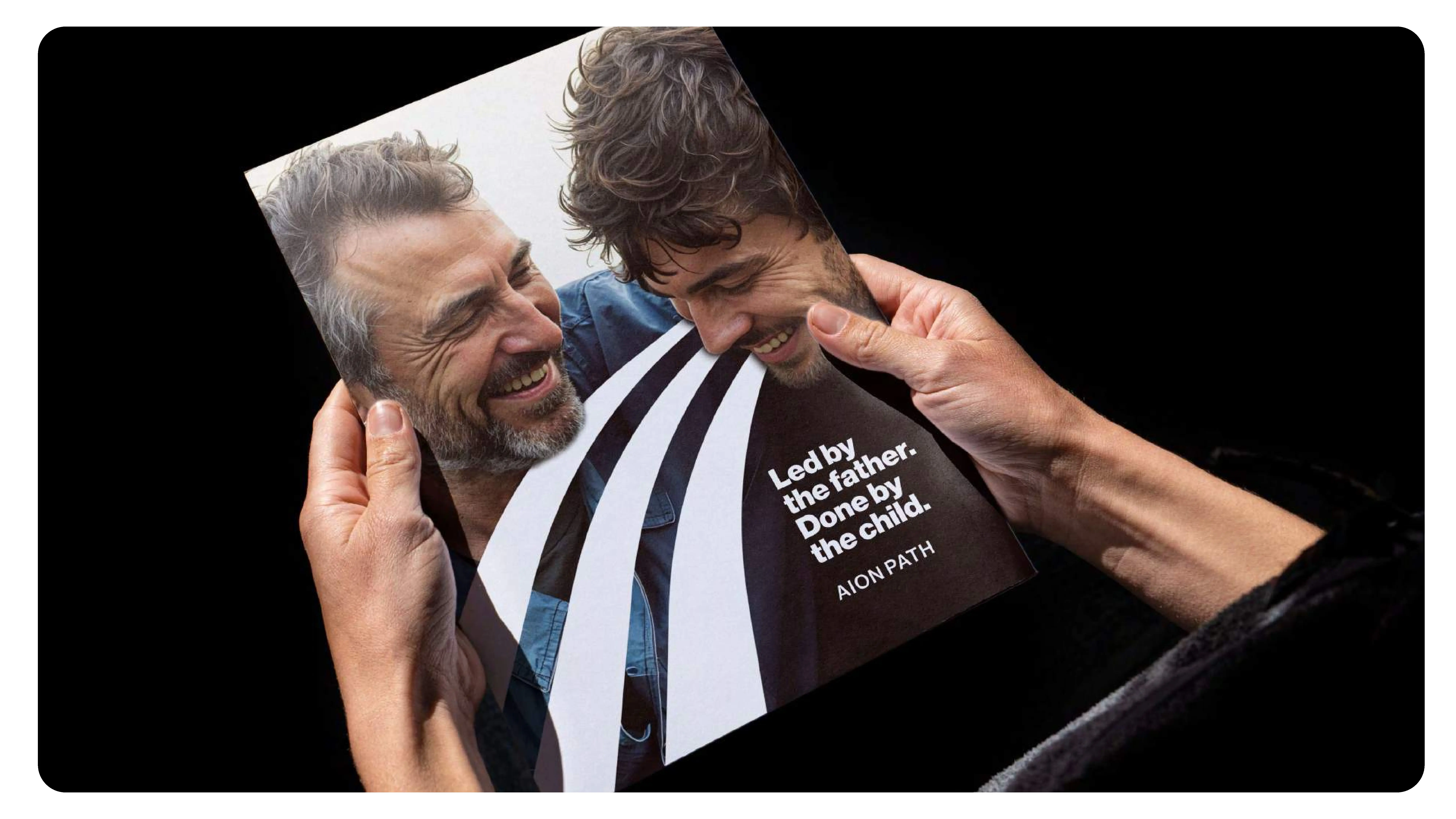 A person holds a brochure with a photo of two men smiling warmly. The text on the cover reads, Led by the father. Done by the child. AIONPATH. Three white stripes partially cover the photo.