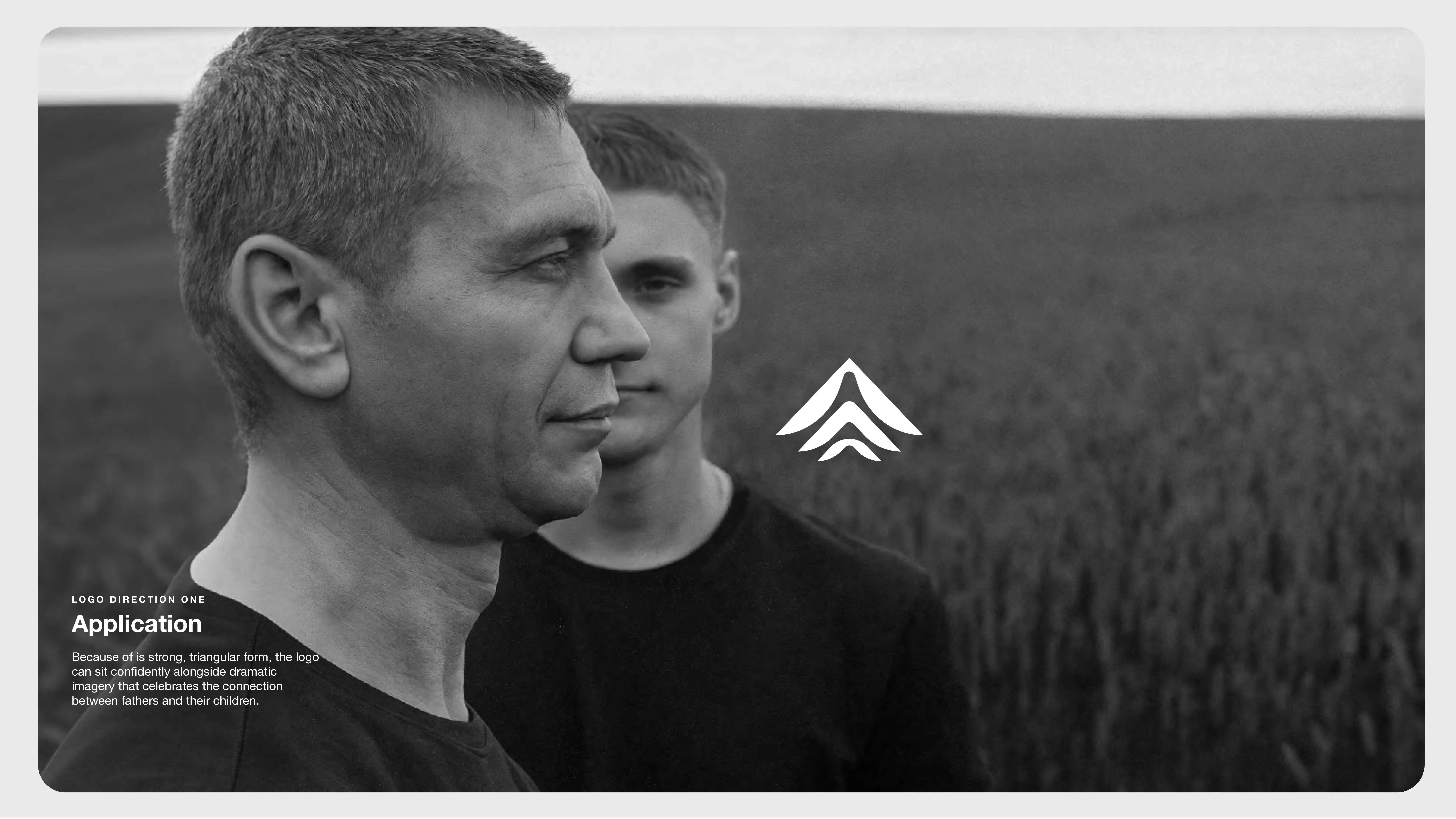 Two men in black shirts stand outside in a field, one in the foreground in profile and one slightly behind him. A white abstract logo is centered over the image. Text appears in the lower left corner.