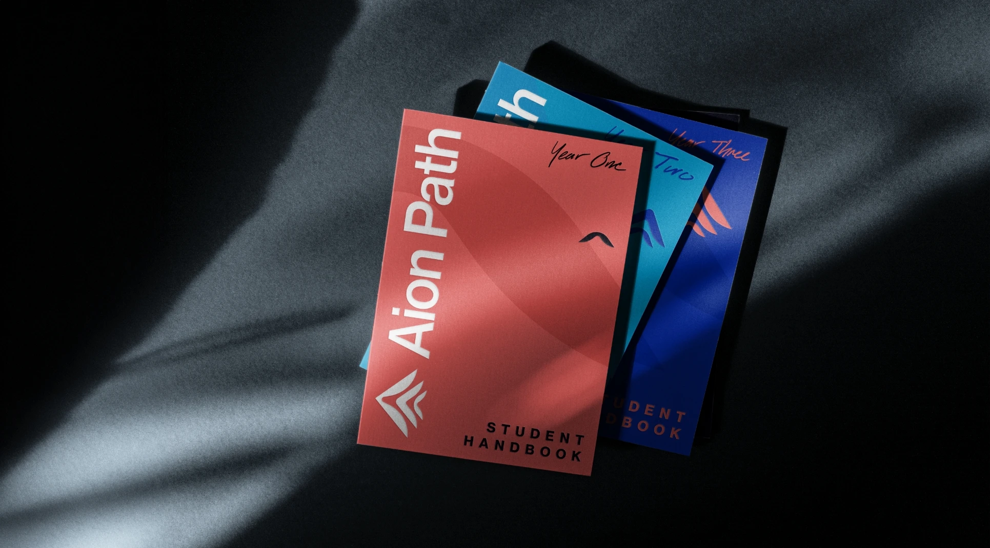 Three student handbooks titled Aion Path are stacked on a dark surface, with the top one in red labeled Student Handbook and two below in blue labeled Year One and Year Two. Sunlight creates diagonal shadows across them.